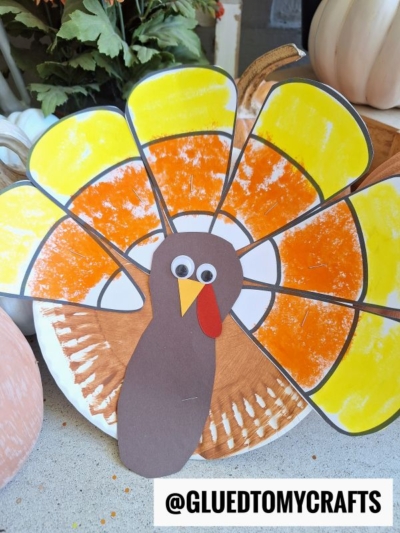 A handmade paper plate candy corn turkey craft with googly eyes, a red wattle, and a fan of orange and yellow tail feathers. The username @GLUEDTOMYCRAFTS is at the bottom of the image.