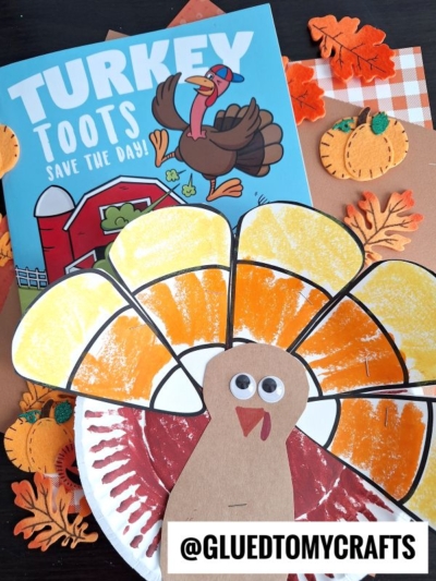 A Thanksgiving-themed craft setup featuring a “Turkey Toots Save the Day!” book, a paper plate candy corn turkey with a painted tail and googly eyes, artificial leaves, and felt pumpkins arranged on a brown surface.