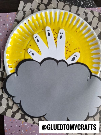 A paper plate painted yellow with a white handprint, ghost faces drawn on fingers, and a gray paper cloud on top, makes a fun Halloween moon craft when set on Halloween-themed paper.