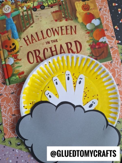 A children’s Halloween moon craft features a yellow paper plate, a white handprint ghost with fun faces, and gray cloud shapes, all set on a festive background with the Halloween in the Orchard book.