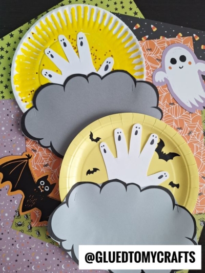 Paper plates with yellow edges create a Halloween moon craft, featuring white handprint ghosts partially hidden by gray clouds. The background showcases Halloween-themed paper with bats and ghosts. A @GLUEDTOMYCRAFTS watermark appears at the bottom.