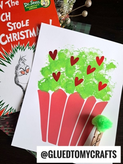 A children’s craft featuring a paper Grinch cupcake with green paint and red hearts, a small clothespin with a pom-pom painted Grinch topper, a Dr. Seuss book, and the Instagram handle @GLUEDTOMYCRAFTS.
