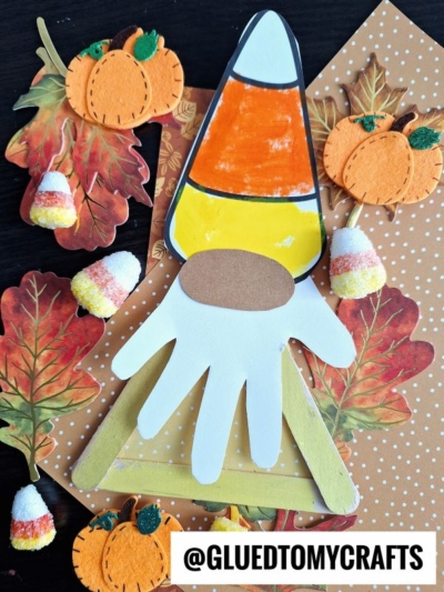 A handmade popsicle stick candy corn gnome with a white paper hand beard is surrounded by felt pumpkins, candy corn, autumn leaves, and the handle @GluedToMyCrafts at the bottom.