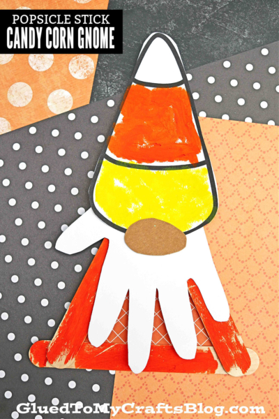 A popsicle stick candy corn gnome with a painted candy corn hat and a white handprint beard sits on orange and black polka dot paper. This whimsical candy corn gnome features a brown nose and playful Halloween colors.