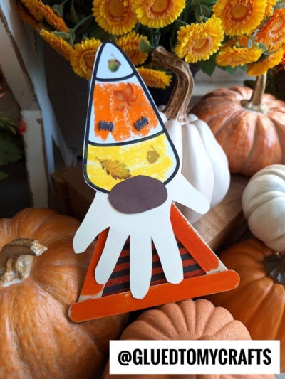 A fall-themed candy corn gnome craft made from paper with a handprint beard, oval nose, and signature candy corn hat, displayed among pumpkins and orange flowers. Text reads @GLUEDTOMYCRAFTS.