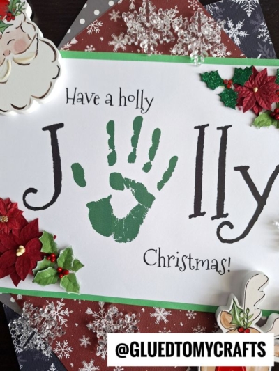 A sign that reads Have a jolly Christmas, featuring a handprint jolly keepsake in place of the letter o, decorated with paper poinsettias, snowflakes, and a Santa cutout.