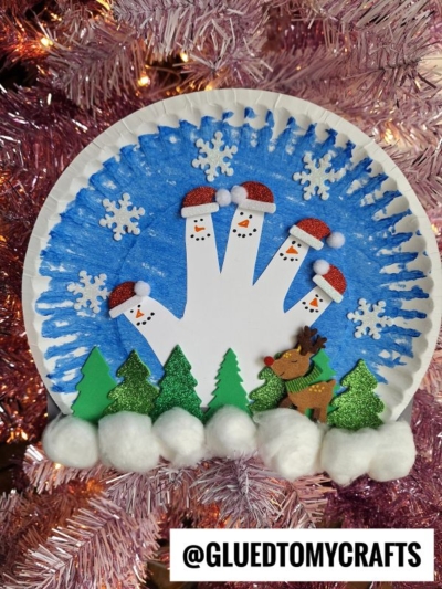 A festive paper plate winter wonderland craft featuring a blue background, a white handprint with snowman faces and Santa hats, snowflake stickers, green trees, cotton snow, a reindeer, and the text @GLUEDTOMYCRAFTS.