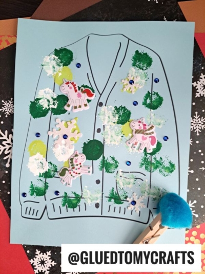 A blue paper with a sweater outline is decorated with paint, sequins, polka dots, stickers of unicorns in scarves, and snowflakes, creating a festive holiday ugly sweater craft. A blue pom-pom paintbrush sits at the bottom.