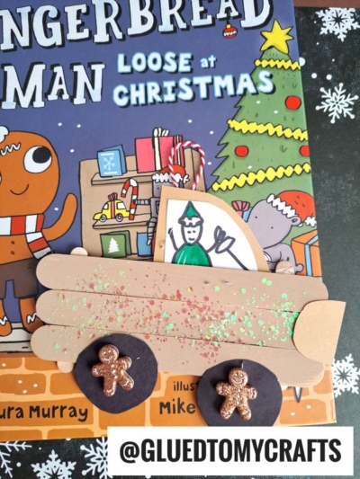 A children’s book titled Gingerbread Man Loose at Christmas sits under a paper craft featuring a popsicle stick gingerbread truck, a drawing of an elf, and two round shapes decorated with mini gingerbread figures.