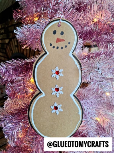 A handmade paper gingerbread snowman ornament with black eyes, an orange nose, and snowflake buttons hangs on a pink Christmas tree. Brown background and text at the bottom read @GLUEDTOMYCRAFTS.