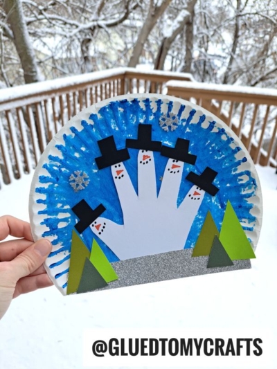 A hand displays a paper plate winter wonderland, painted blue with white handprint snowmen in black hats. Green trees and glittery snowflakes add festive charm. Snowy trees and a wooden railing complete the cozy background scene.