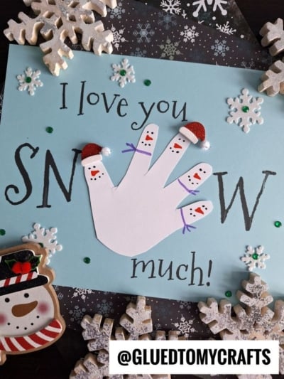 A handmade card with a white handprint decorated as snowmen wearing red Santa hats, reading I love you snow much! Surrounded by sparkling snowflakes, sequins, and a cute snowman cookie.