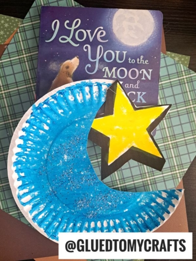 A paper plate moon painted blue with glitter forms a crescent shape, next to a yellow star. Behind them is the book I Love You to the Moon and Back. Green plaid paper and a brown surface are under the craft.