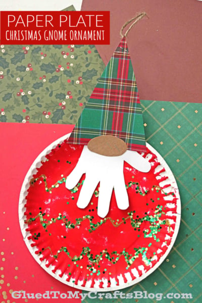 A Christmas gnome ornament made from a paper plate painted red, with green glitter, a white paper handprint beard, plaid paper hat, and brown oval nose. Perfect for gnome Christmas tree ornaments. Festive paper backgrounds. Text: Paper Plate Christmas Gnome Ornament.