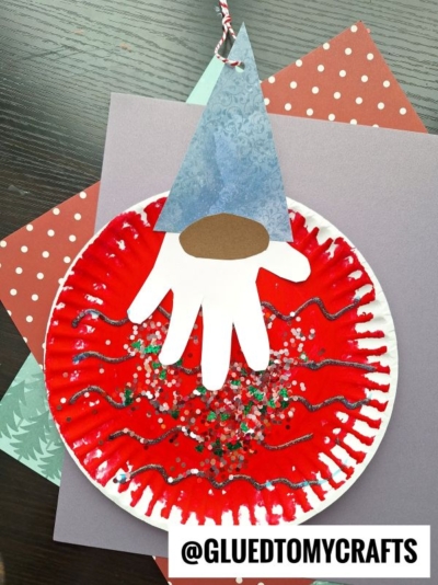 A festive craft resembling a holiday gnome, this red paper plate is decorated with glitter, paint, and a cut-out white handprint—perfect for making charming gnome Christmas ornaments. Craft materials are in the background.