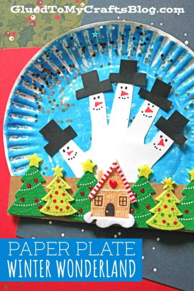 A paper plate winter wonderland craft features a painted paper plate winter scene with handprint snowmen sporting black hats. Felt trees and a gingerbread house decorate the snowy landscape. Text reads Paper Plate Winter Wonderland.
