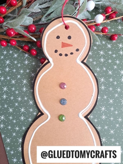 A handmade paper snowman gingerbread ornament made from brown paper with a black outline, decorated with three glittery buttons and a drawn face, lies on green star-patterned paper with red and white berry sprigs.