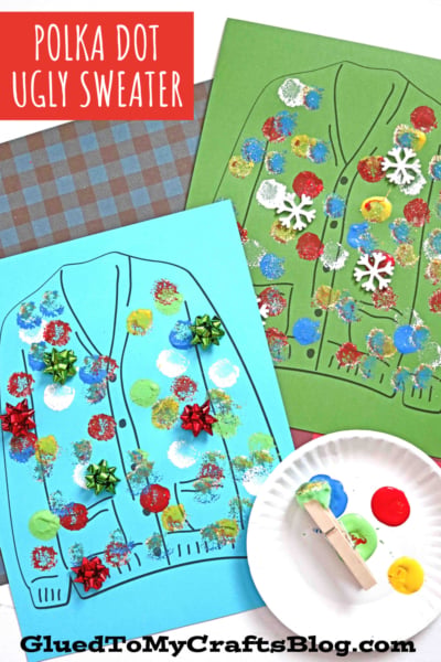 Two pieces of paper, one blue and one green, each featuring a polka dot ugly sweater outlined and decorated with colorful paint dots, glitter, bows, and snowflake cutouts. A plate with paint and a clothespin are nearby.