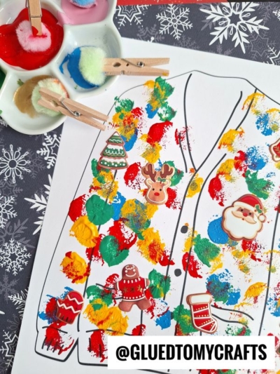 A Christmas polka dot ugly sweater coloring page is decorated with multicolored paint blobs, stickers of Santa, reindeer, and treats. Clothespins with pom poms and a paint palette are nearby. Snowflake patterns form the background.