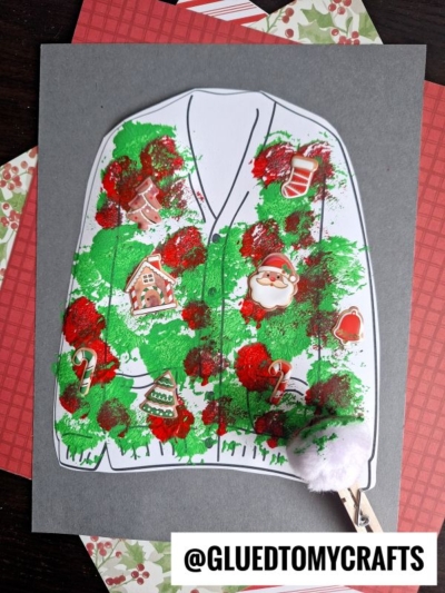 A polka dot ugly sweater craft on gray paper, decorated with green and red paint, holiday stickers, and a white pom-pom. Festive patterned papers are in the background.