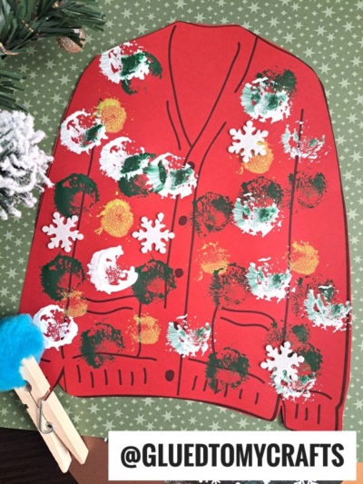 A red paper cutout of a polka dot ugly sweater, decorated with green, white, and gold spots and white snowflake shapes, is displayed on a green patterned background. @GLUEDTOMYCRAFTS is labeled at the bottom.
