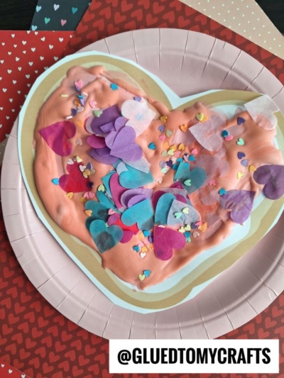A paper plate craft featuring a large heart shape covered in pink and white paint, resembling a puffy paint sugar cookie, decorated with colorful tissue paper hearts and heart-shaped confetti, displayed on heart-patterned paper. Text reads “@GLUEDTOMYCRAFTS.”.
