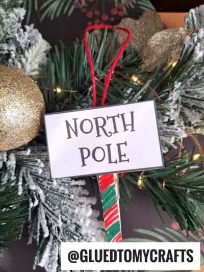 A festive clothespin North Pole ornament hangs on a Christmas tree, adorned with a glittery gold bauble, faux snow, and green branches. The handle is wrapped in red and green yarn. @GLUEDTOMYCRAFTS is at the bottom.
