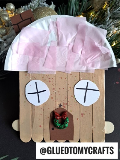 A gingerbread face made from craft sticks, with pink tissue paper on top, paper eyes with Xs, a green and red glitter nose, and a brown paper mouth—perfect as a gingerbread house ornament. Holiday decorations and pine branches are in the background.