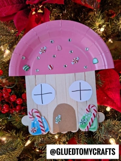 A pink paper plate and craft sticks form a gingerbread house ornament, decorated with paper candies, jewels, and candy canes, hanging on a Christmas tree with red berries and poinsettias. @GLUEDTOMYCRAFTS text at bottom.