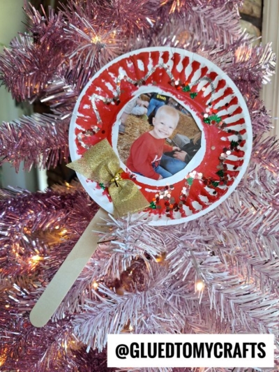 A lollipop photo ornament made from a paper plate with a child’s photo, red and white paint, glitter, a gold bow, and a craft stick hangs on a pink Christmas tree.