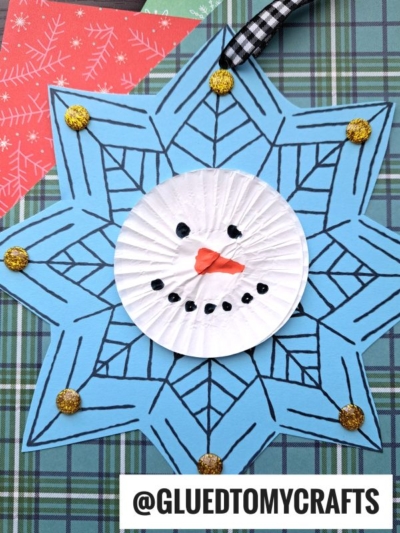 A snowman snowflake ornament features a blue star with black lines, gold glitter dots, and a white cupcake liner snowman face with a carrot nose and smile, all displayed on a plaid background.