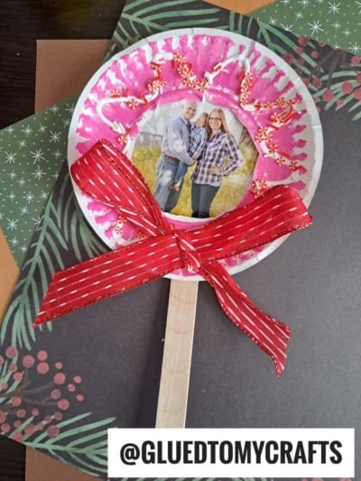 A handmade lollipop photo ornament featuring a couples photo in the center, with a red bow, wooden stick handle, and pink and white decorations on a paper plate, displayed on festive paper backgrounds.
