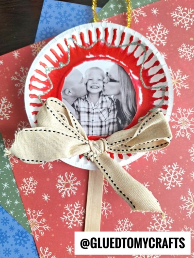 A handmade lollipop photo ornament features a photo of a smiling child being kissed on both cheeks, framed by a red-painted paper plate edge, with a beige bow below and festive snowflake-patterned paper in the background.