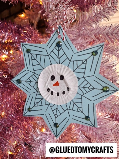 A handmade snowman snowflake ornament with a snowman face in the center, crafted from blue paper and a white cupcake liner, hangs on a pink Christmas tree. The username @GLUEDTOMYCRAFTS appears at the bottom.