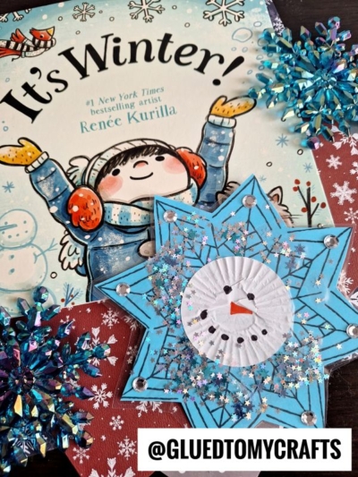 A children’s book titled It’s Winter! by Renée Kurilla is surrounded by blue and silver snowflake ornaments and a handcrafted snowman on a blue star. The @gluedtomycrafts handle is at the bottom.