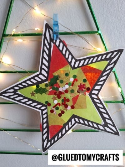 A colorful, handmade stained glass Christmas star ornament decorated with tissue paper, sequins, and confetti hangs from a blue clip on a green wire Christmas tree with string lights. Text reads “@GLUEDTOMYCRAFTS” at the bottom.