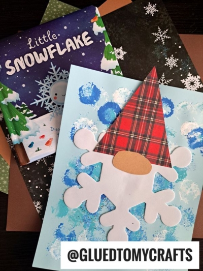 A children’s craft project featuring a craft foam snowflake gnome made from paper rests on blue painted paper, beside the book Little Snowflake and decorative sheets. @GLUEDTOMYCRAFTS is labeled at the bottom.