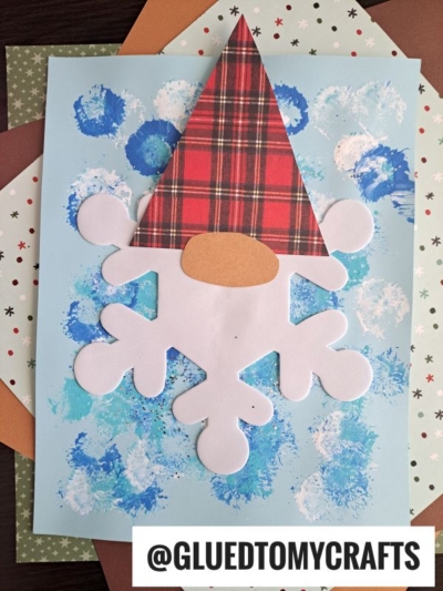 A handmade craft foam snowflake gnome features a plaid hat, oval nose, and white snowflake-shaped beard on a blue and white painted background. Various patterned papers are layered beneath. Text reads @GLUETOMYCRAFTS.