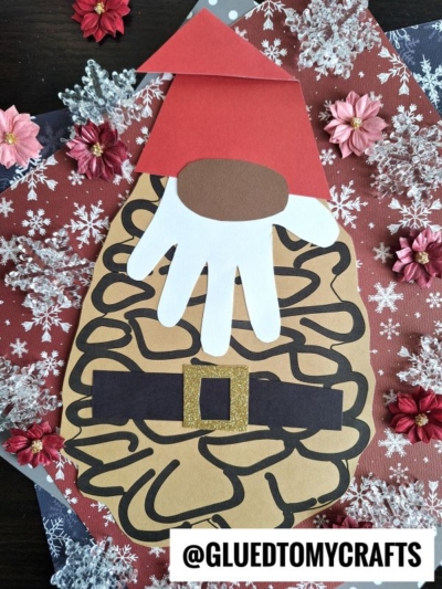 A pinecone Santa gnome with a handprint beard, gold-buckled belt, and festive hat sits among snowflakes and pink flowers on a cheerful background.