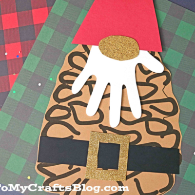 A paper craft pinecone santa gnome with a brown, pinecone-shaped body, red hat, white paper hand-beard, and black belt with gold buckle. Plaid background sets a festive scene.