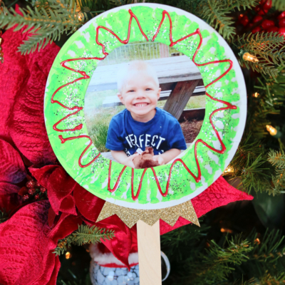 A handmade lollipop photo ornament featuring a child's picture hangs on the Christmas tree. Decorated with green and red patterns and two craft sticks as the handle, it’s surrounded by red ribbon and festive foliage.