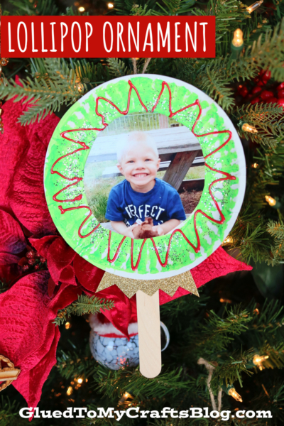 A handmade lollipop photo ornament featuring a child's picture hangs on the Christmas tree. Decorated with green and red patterns and two craft sticks as the handle, it’s surrounded by red ribbon and festive foliage.