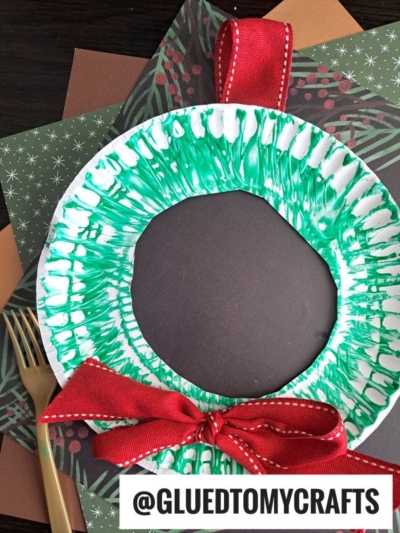 A paper plate fork painted green to resemble a Christmas wreath, with a red ribbon bow at the bottom and a loop at the top, placed on festive papers; @GLUEDTOMYCRAFTS is written at the bottom.