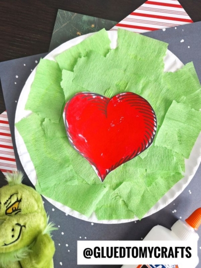A paper plate Grinch heart craft features a large red heart in the center, surrounded by torn green tissue paper. A Grinch plush toy, glue bottle, and the handle @GLUEDTOMYCRAFTS are visible nearby.