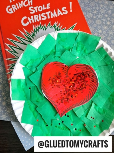 A festive paper plate Grinch heart craft inspired by How the Grinch Stole Christmas, made with green tissue squares and a bold red heart. The beloved book peeks out in the background. Text: @GLUEDTOMYCRAFTS.