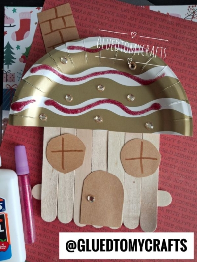 Craft of a gingerbread face made from craft sticks, paper, felt, and sequins, with a gold and red hat, glue, and marker nearby—reminiscent of a whimsical gingerbread house ornament; background features red paper with festive text.