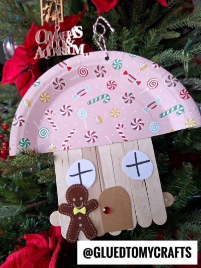 A gingerbread house ornament, handmade from a decorated paper plate, craft sticks, and paper shapes, hangs on a Christmas tree. Red poinsettias and a silver sign are in the background. @GLUEDTOMYCRAFTS is written at the bottom.