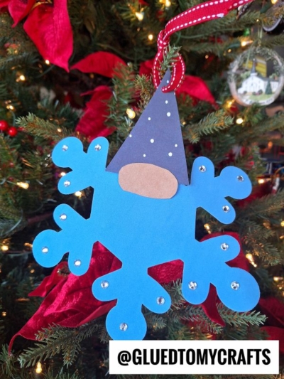 A blue craft foam snowflake gnome ornament with a gnome face and hat, decorated with clear gems, hangs on a Christmas tree with red ribbon and ornaments. Text at the bottom reads @GLUEDTOMYCRAFTS.