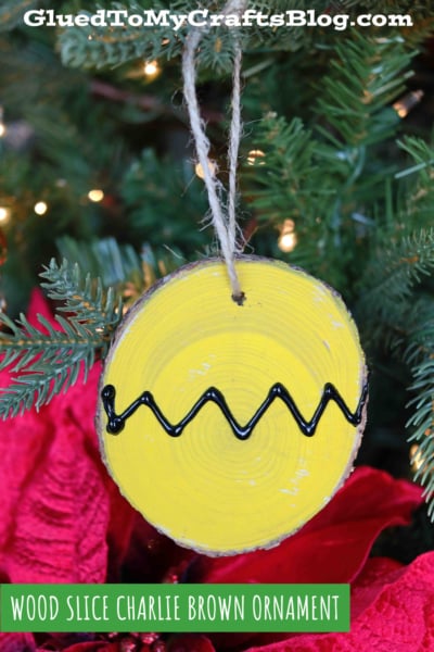 A round wood slice Charlie Brown ornament, painted yellow with a black zigzag line, hangs on a Christmas tree beside red poinsettia flowers. Text reads “WOOD SLICE CHARLIE BROWN ORNAMENT.”.