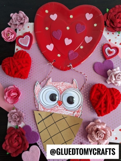 A Valentine’s Day craft featuring a paper owl with large googly eyes in a brown cone, a heart-shaped red balloon like a paper plate hot air balloon, and decorative pink and red flowers and hearts on a pink polka dot background. @GLUEDTOMYCRAFTS is written below.